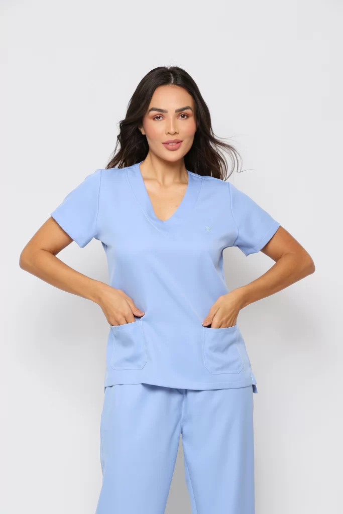 Modelo vestida de scrub hospitalar. O uniforme consiste em uma blusa de manga curta com decote em V e dois bolsos frontais, e uma calça combinando. O scrub é ideal para clínica, hospital.